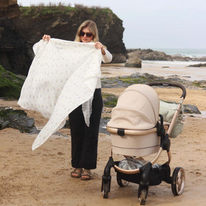 Avery Row Coastline Muslin Swaddle shown in lifestyle photo on beach with mother and baby.