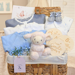 Baby boy hamper basket with presents including organic clothing in blue and white with a lamb toy and milestone discs.