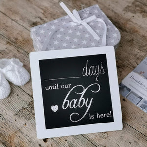 White-framed baby countdown plaque resting on a wooden surface beside a grey star-patterned cloth.
