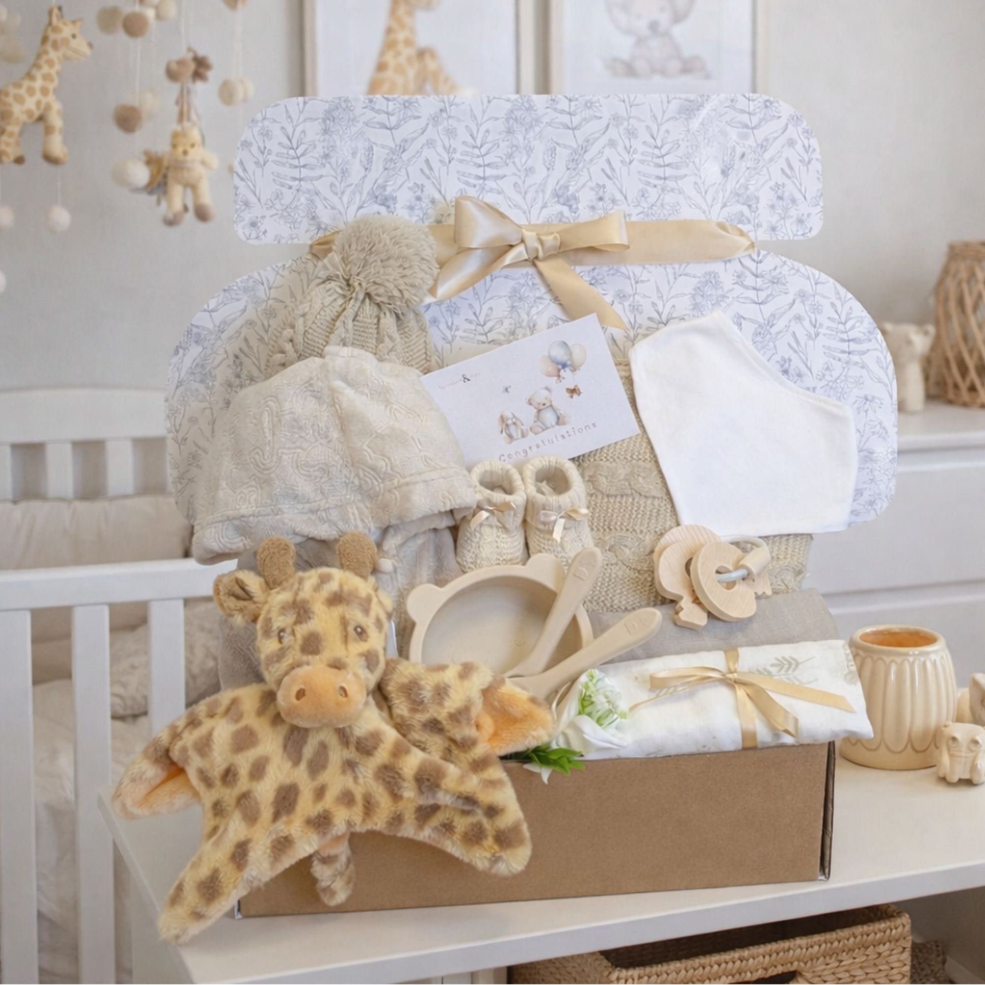 Newborn hamper gift box with gifts including a giraffe baby comforter and beige knit blanket and feeding essentials.