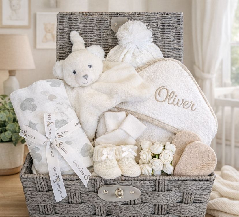 A baby hamper basket with white presents including a personalised bath towel and bear comforter.