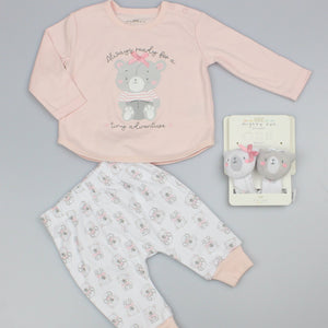 Baby girl clothes set laid flat on grey: pink long-sleeved bear top, white printed trousers and two wrist rattles.