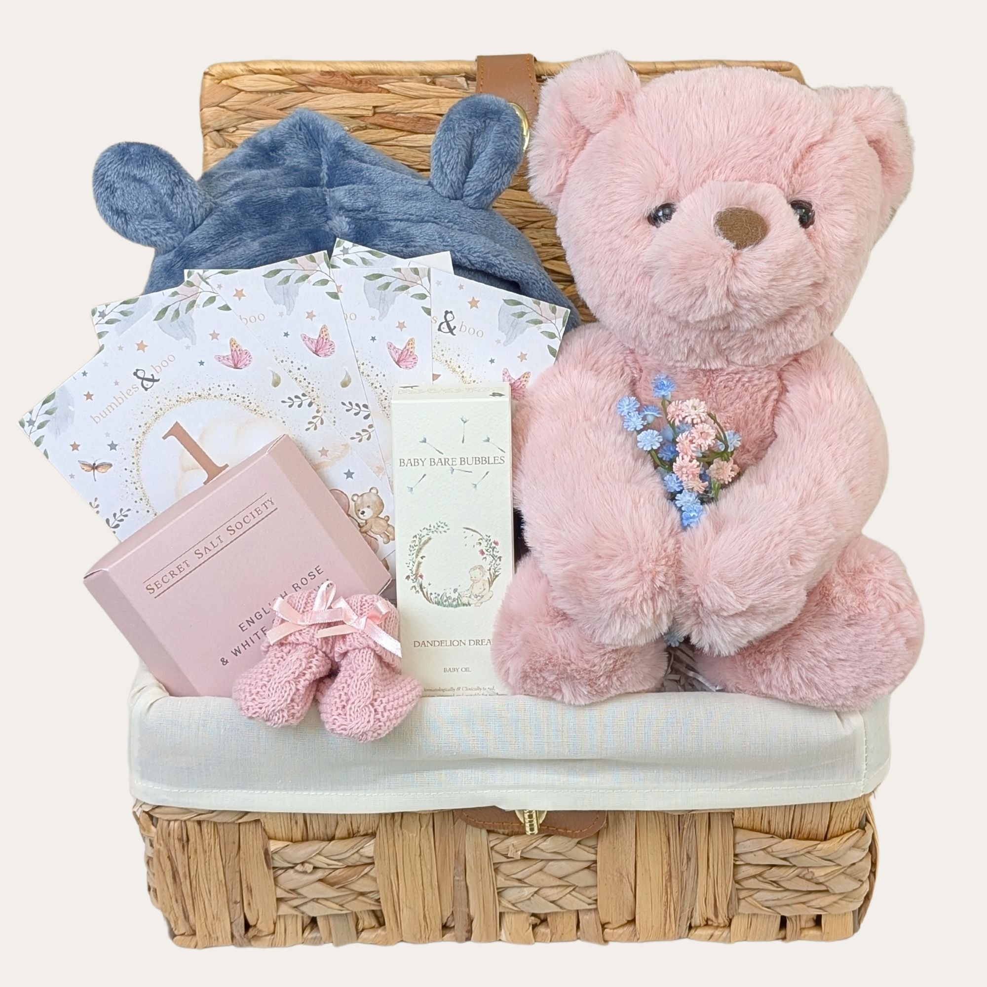 Baby girl gifts hamper basket containing a dusty pink teddy bear, blue baby dressing gown and pink baby presents in a seagrass keepsake basket, with other baby essentials.