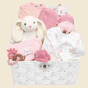 Luxury baby girl gifts in a hamper basket. Presents include white and pink bunny-themed baby clothes and white rabbit soft toy.
