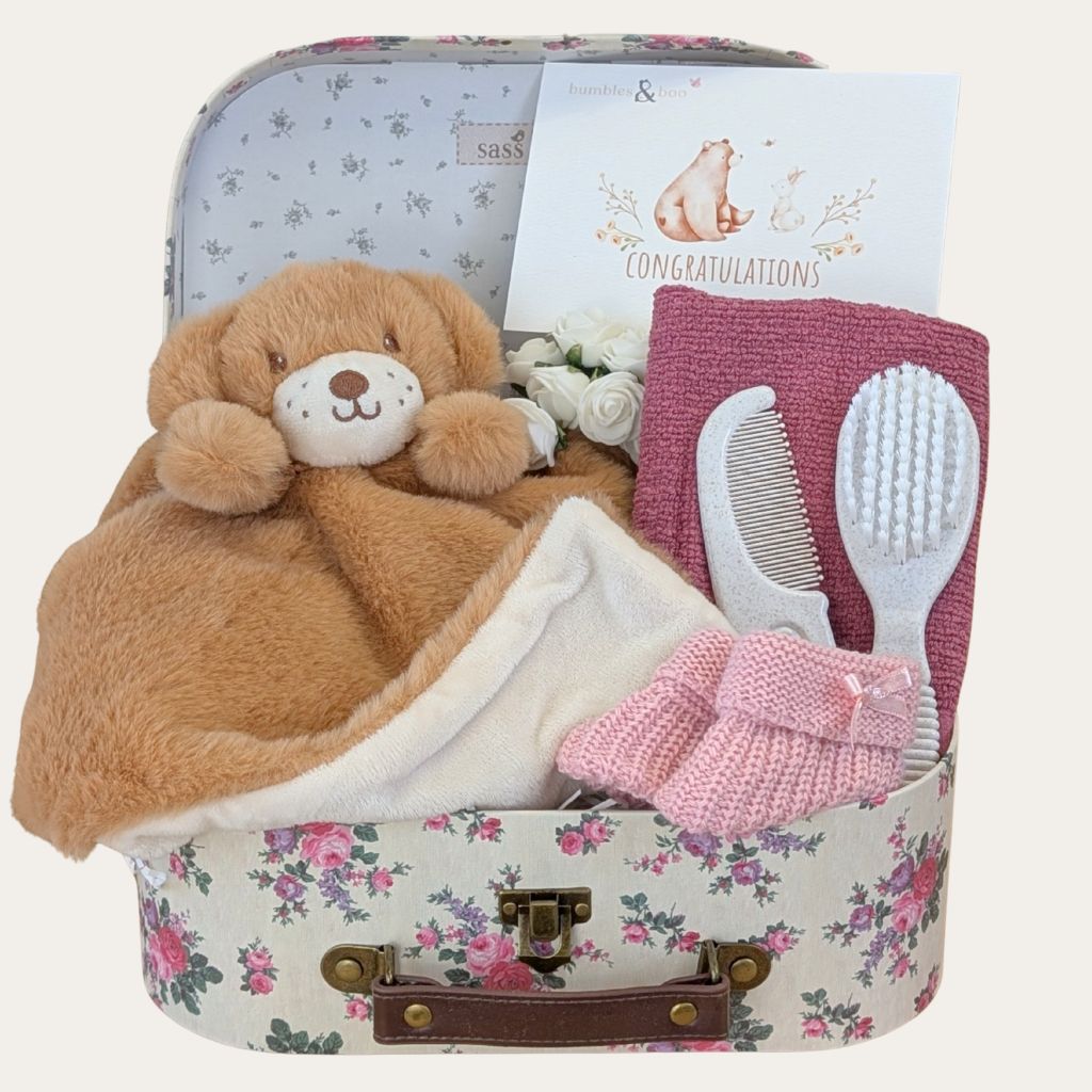Baby girl hamper with gifts in a floral-patterned suitcase, teddy bear, baby items and a card.