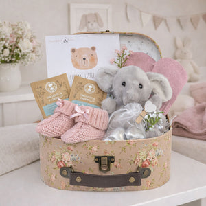A floral gift basket with presents for a baby girl including elephant and mittens.