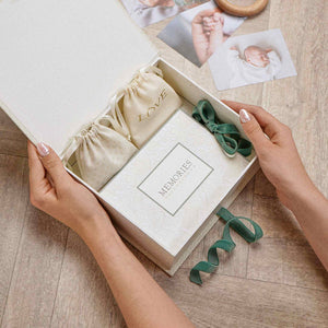 Hands holding an open baby keepsake box containing drawstring pouches and a ribbon-tied ‘MEMORIES’ item.