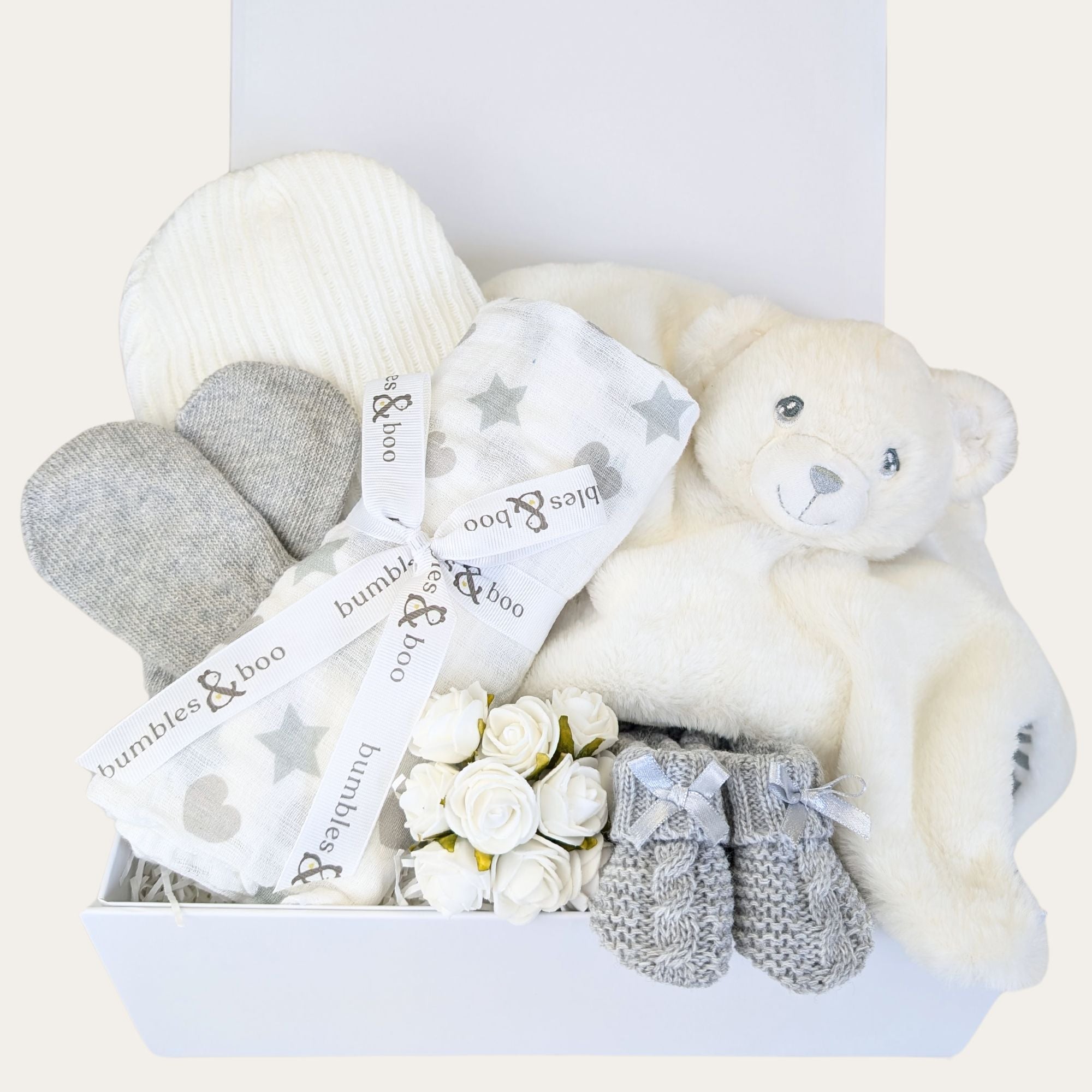 A baby shower gifts hamper containing a teddy bear, a knitted hat, mittens, and booties in a neutral color palette, presented in a white box with a blank greeting card.