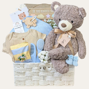 Baby shower gifts in a white hamper basket with blue and brown presents including teddy bear.