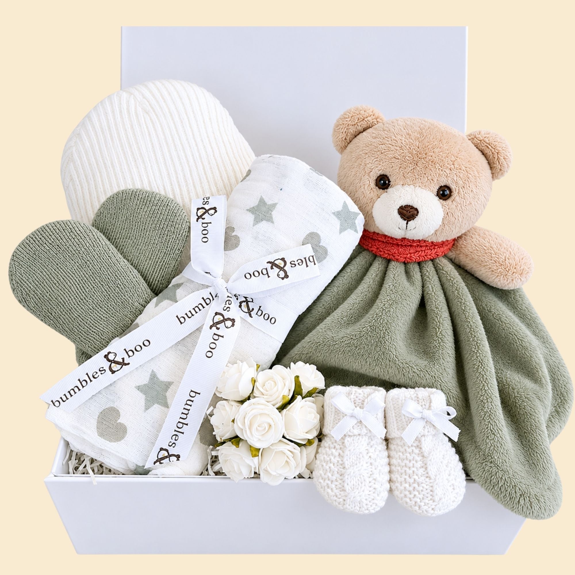 A baby shower gifts hamper containing a teddy bear, a knitted hat, mittens, and booties in a neutral color palette, presented in a white box with a blank greeting card.