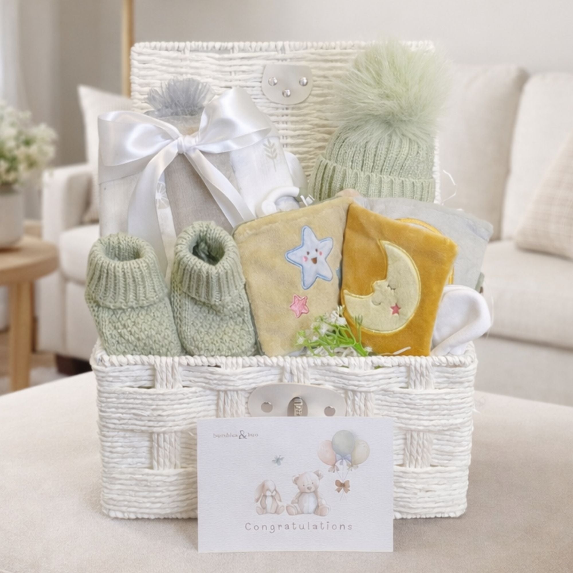 Baby shower hamper with gifts including sage green knit hat and mittens and a baby book in a white wicker basket.