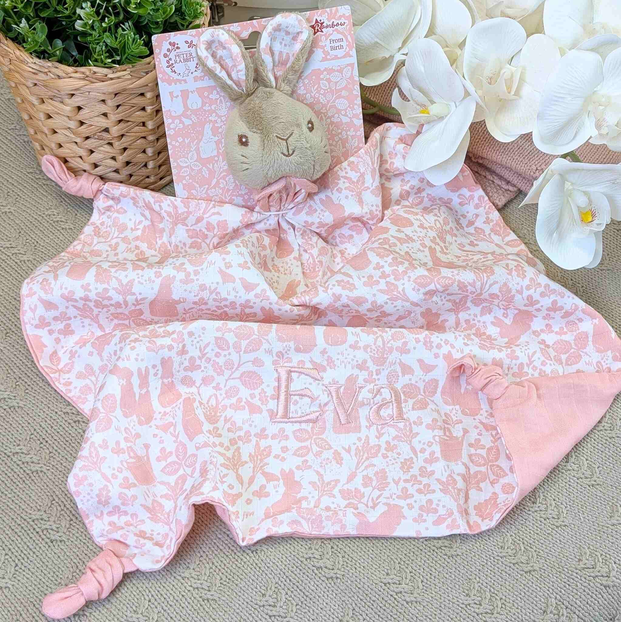 Pink floral blanket with 'Eva' embroidered, bunny plush toy, and flowers on a textured surface.