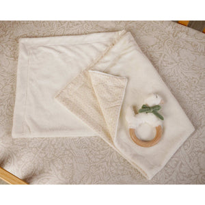 Cream baby blanket with a folded patterned corner and goose ring teether on a patterned surface.