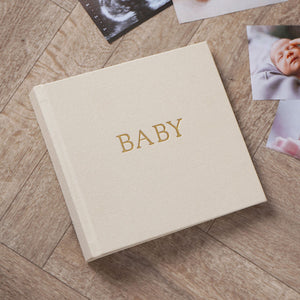 Neutral linen baby photo album with gold BABY lettering beside photographs on a wooden surface