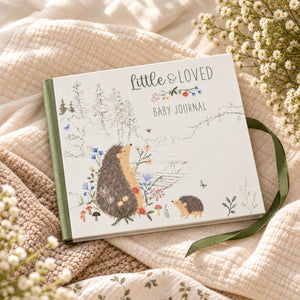 A baby journal titled 'Little & Loved Baby Journal' with a white cover, illustrated hedgehogs, delicate floral details, and a green ribbon bookmark