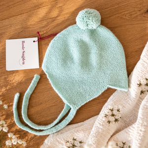 Mint green cashmere baby bonnet with a pom-pom, ear flaps and ties on a wooden tabletop.