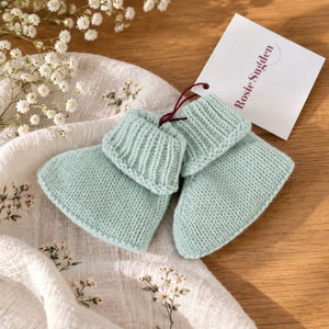 Pair of mint green cashmere baby booties with ribbed cuffs beside a cream blanket and white flowers.