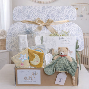 New baby gifts in a hamper box with a baby journal and teddy bear comforter.