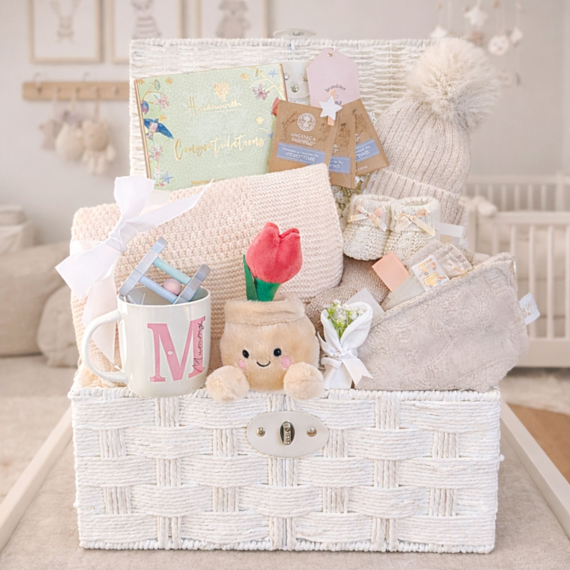 New mum gifts hamper packed with presents for mum and baby in a white wicker basket featuring a knitted baby blanket, soft toy, organic baby products and congratulations card.