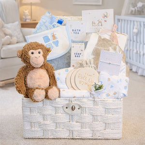 Newborn baby boy gifts in a white hamper basket which includes a steiff monkey soft toy and blue and white clothing set.