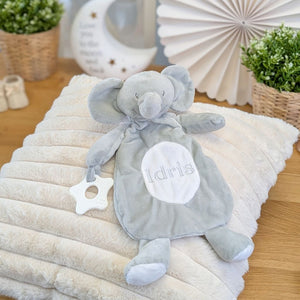Gray plush elephant toy with 'Idris' on a white pillow, surrounded by decorative items.