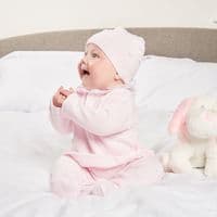 Baby sitting on a white blanket wearing the pink bobble knit top, leggings and matching hat.