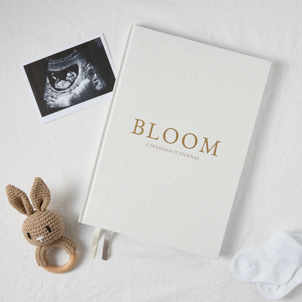 Pregnancy journal titled 'BLOOM' with an ultrasound photo and a crocheted rabbit rattle on a light background
