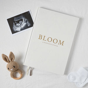 Pregnancy journal titled 'BLOOM' with an ultrasound photo and a crocheted rabbit rattle on a light background
