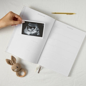 Hand holding an ultrasound photo over an open notebook with a crocheted rabbit toy on a light surface.