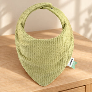 Lemongrass green textured bandana bib resting on a wooden dresser beside a softly lit cream wall.