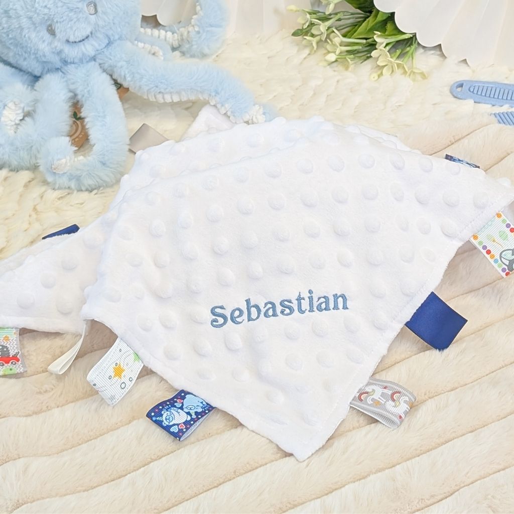 Personalised white baby comforter with blue taggie ribbons, embroidered with baby's name.