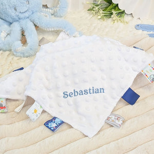 Personalised white baby comforter with blue taggie ribbons, embroidered with baby's name.
