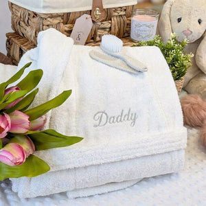 Folded white Daddy dressing gown with embroidered lettering beside flowers and a gift basket.