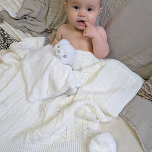 White cable knit baby blanket with pom poms, with a baby sitting underneath it.