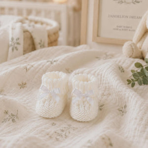 Pair of pale knitted baby booties with bows on a floral blanket.