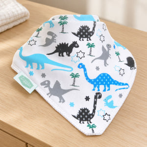 White Ziggle bandana bib with blue, grey and black dinosaurs and green trees displayed on a wooden nursery dresser.