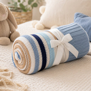 A rolled knit baby blanket with horizontal stripes in light blue, navy, beige, and white, tied with a white ribbon bow