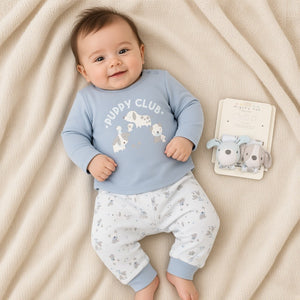 Baby wearing blue Puppy Club top and white puppy-print trousers, with two wrist rattles on a cream blanket.