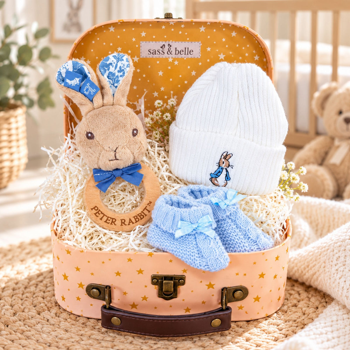 A baby boy gift set arranged inside a pastel suitcase with gold stars. The set includes a plush Peter Rabbit teething ring