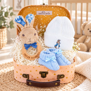 A baby boy gift set arranged inside a pastel suitcase with gold stars. The set includes a plush Peter Rabbit teething ring