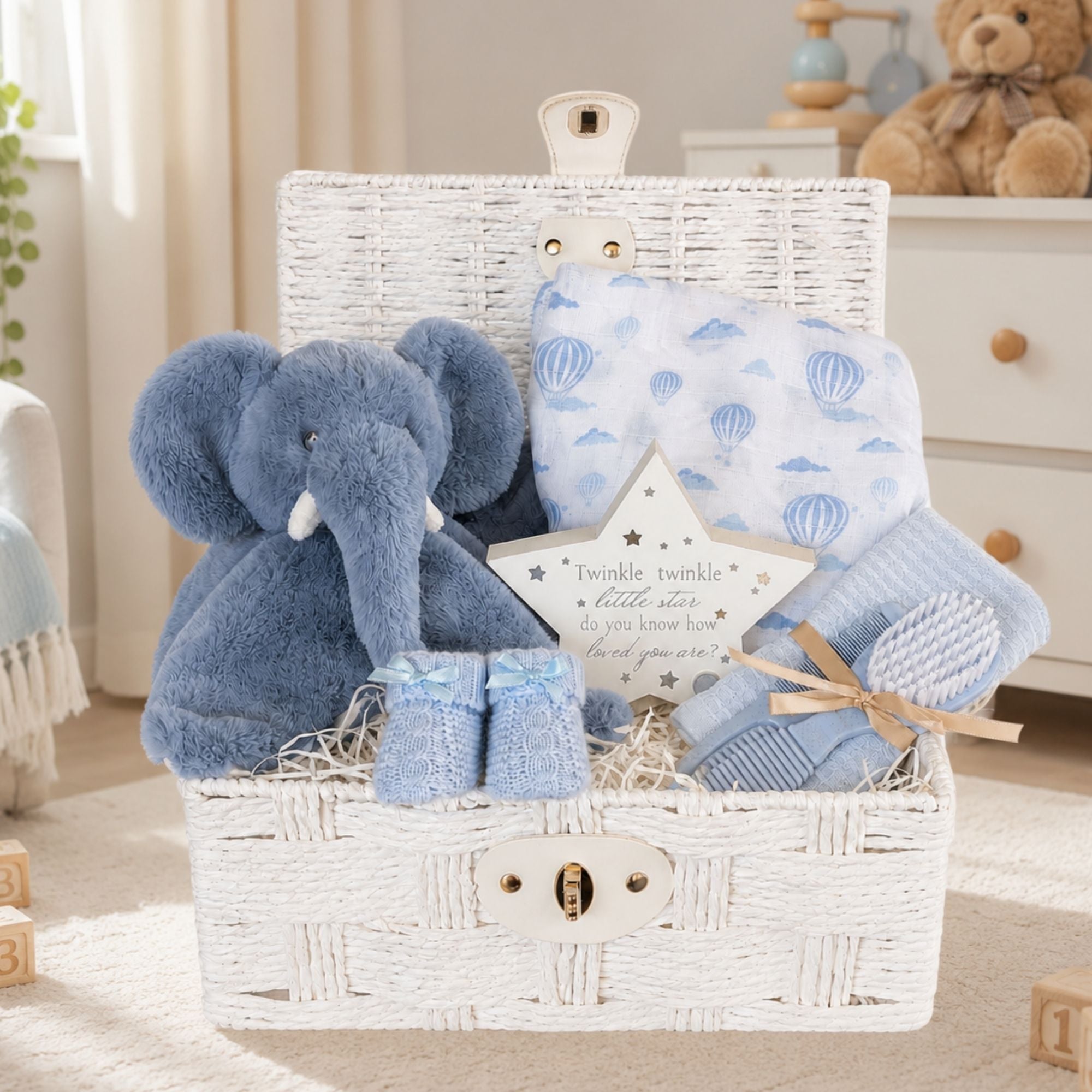 A baby boy gift hamper set in a white wicker basket, containing a blue plush elephant toy and light blue baby boy booties