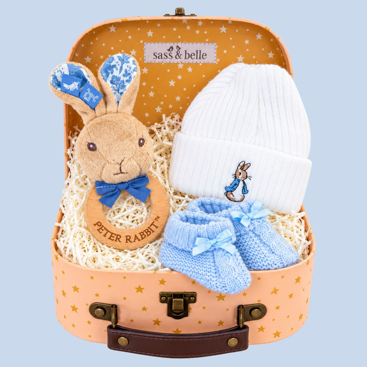 A baby boy gift set arranged inside a pastel suitcase with gold stars. The set includes a plush Peter Rabbit teething ring