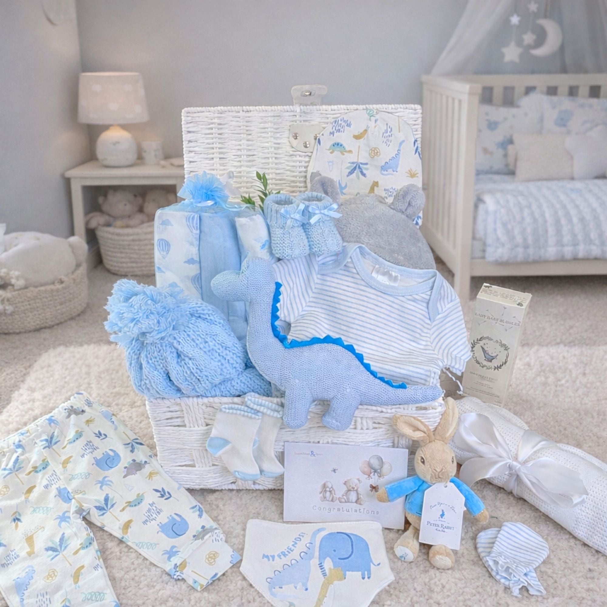 Baby boy gifts set in a white hamper basket with blue organic dinosaur rattle and dinosaur baby clothing.