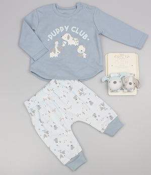 Baby boy clothes laid flat on grey: blue Puppy Club top, white printed trousers and matching wrist rattles.