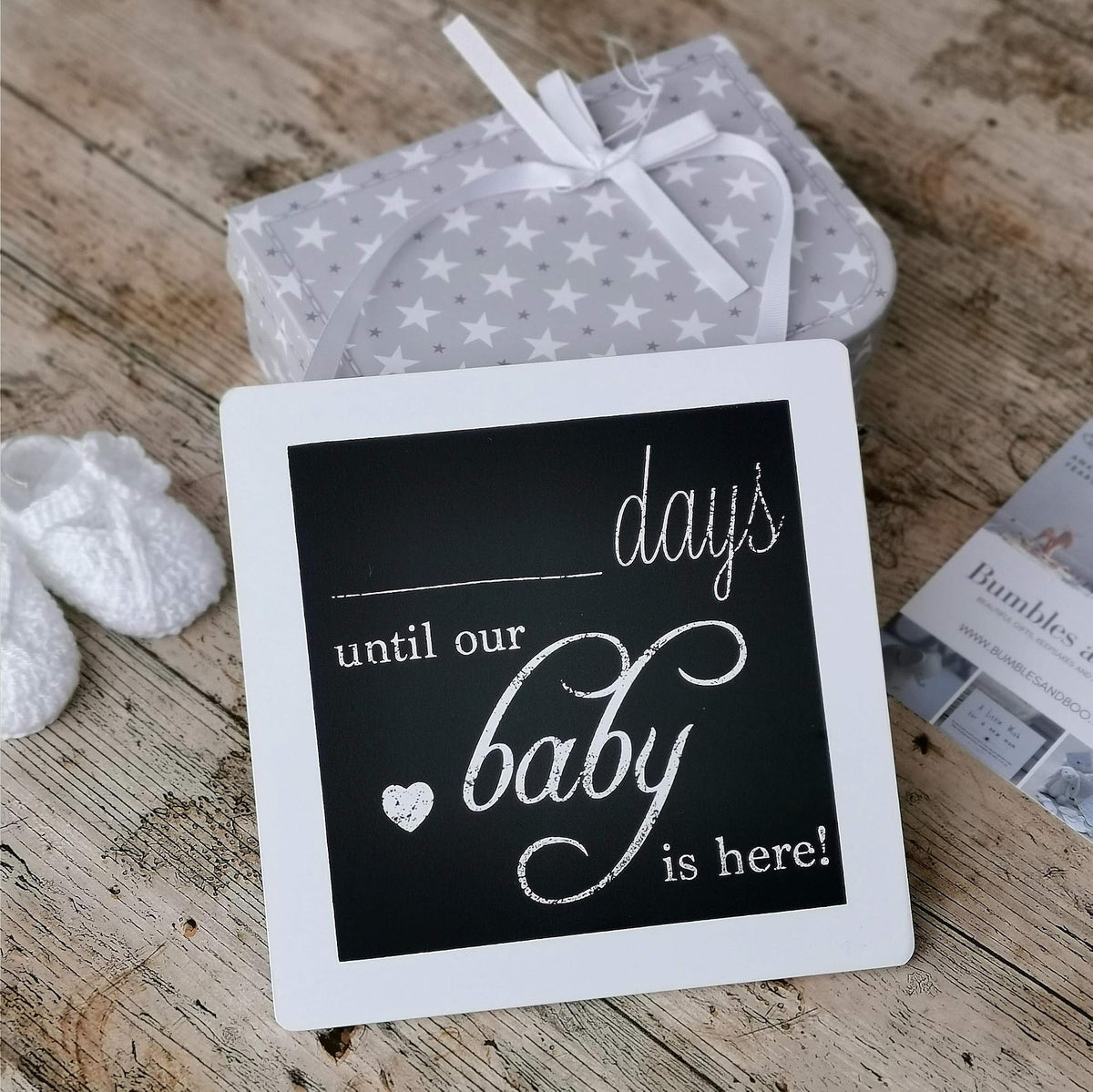 White-framed baby countdown plaque resting on a wooden surface beside a grey star-patterned cloth.