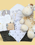 Baby gifts in a wicker hamper basket, including giraffe soft toy, milestone cards and white towel.