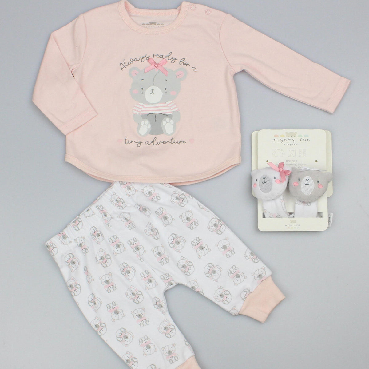Baby girl clothes set laid flat on grey: pink long-sleeved bear top, white printed trousers and two wrist rattles.