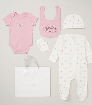 A 5-piece baby girls clothing gift set including a pink bodysuit, a white sleepsuit with pink hearts, a matching hat, a bib, and a pair of mittens, all presented on a white background.