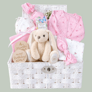 Newborn baby girl gifts in a white hamper basket with organic pink baby clothing and bunny soft toy.