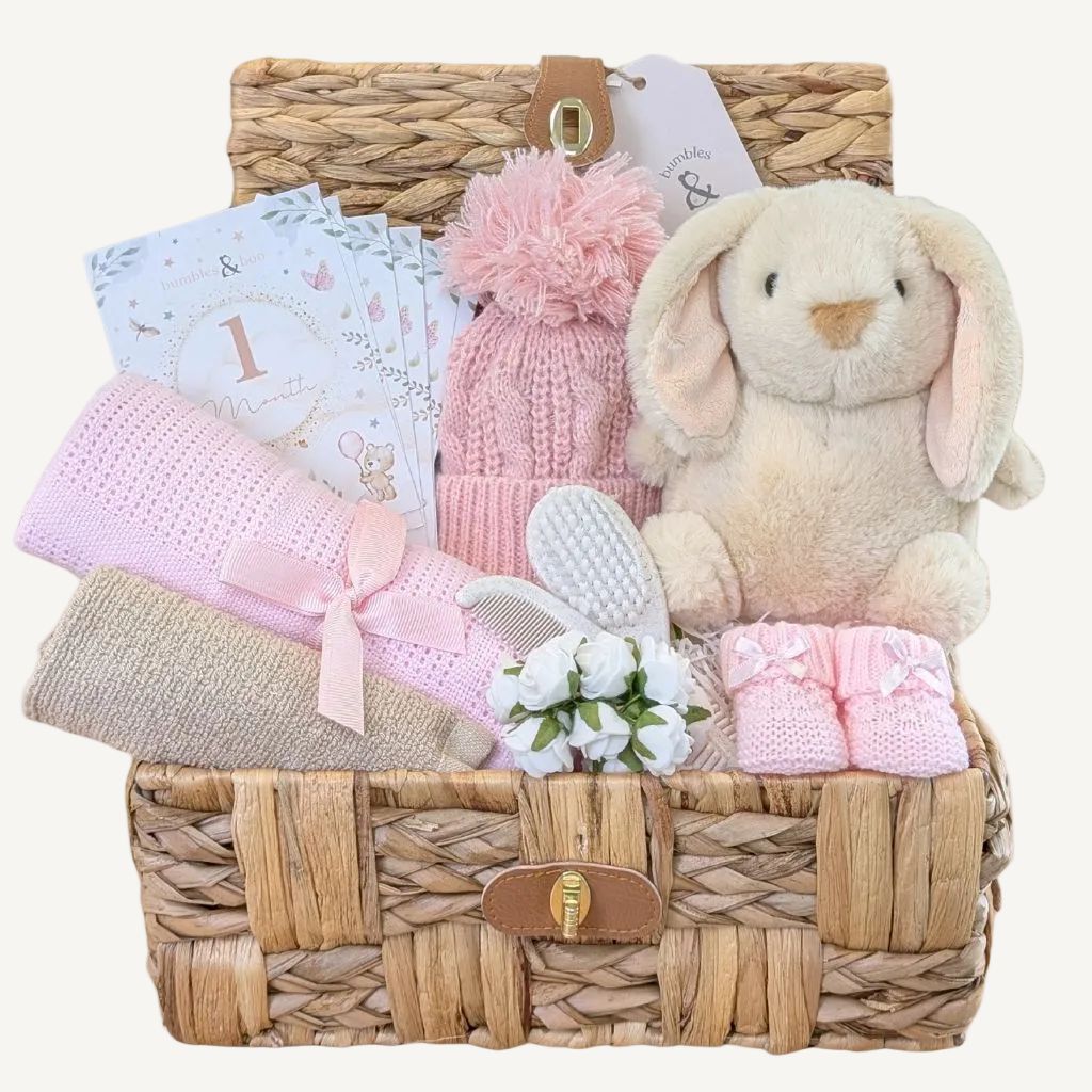 Baby girl gifts hamper with presents including plush bunny toy, blanket and booties in a woven basket.